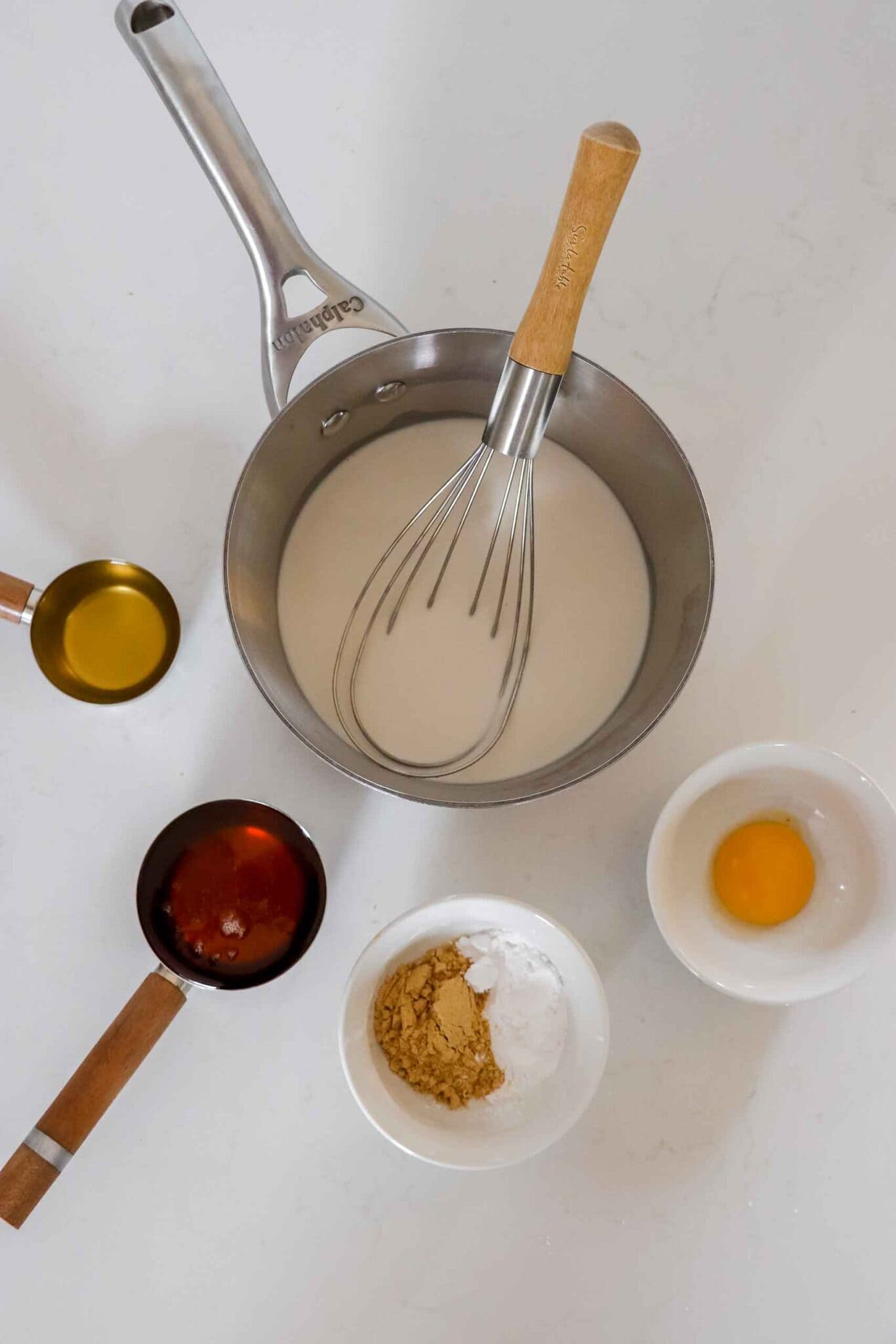 Paleo Mustard Sauce in a clear mason jar on a wood board. Behind it is dry mustard in a copper measuring spoon, cup of Apple cider vinegar, an egg and a cut off jar of milk. www.atwistedplate.com/paleo-mustard-sauce/