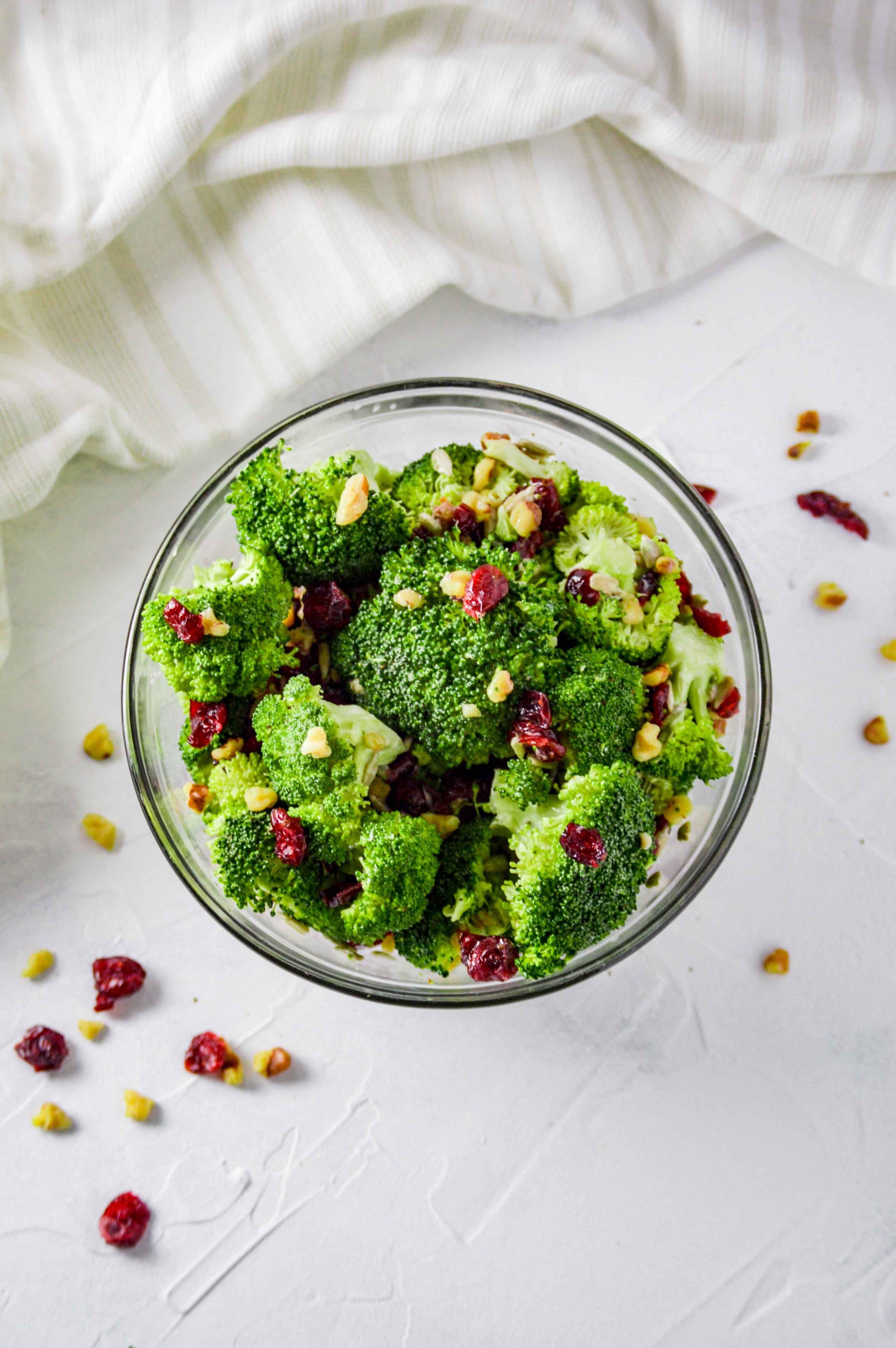 Top view of Broccoli salad in a clear bowl against a white background.  there are walnuts and dried cranberries scattered around and a white towel at the top of the image.  https://www.atwistedplate.com/broccoli-salad/