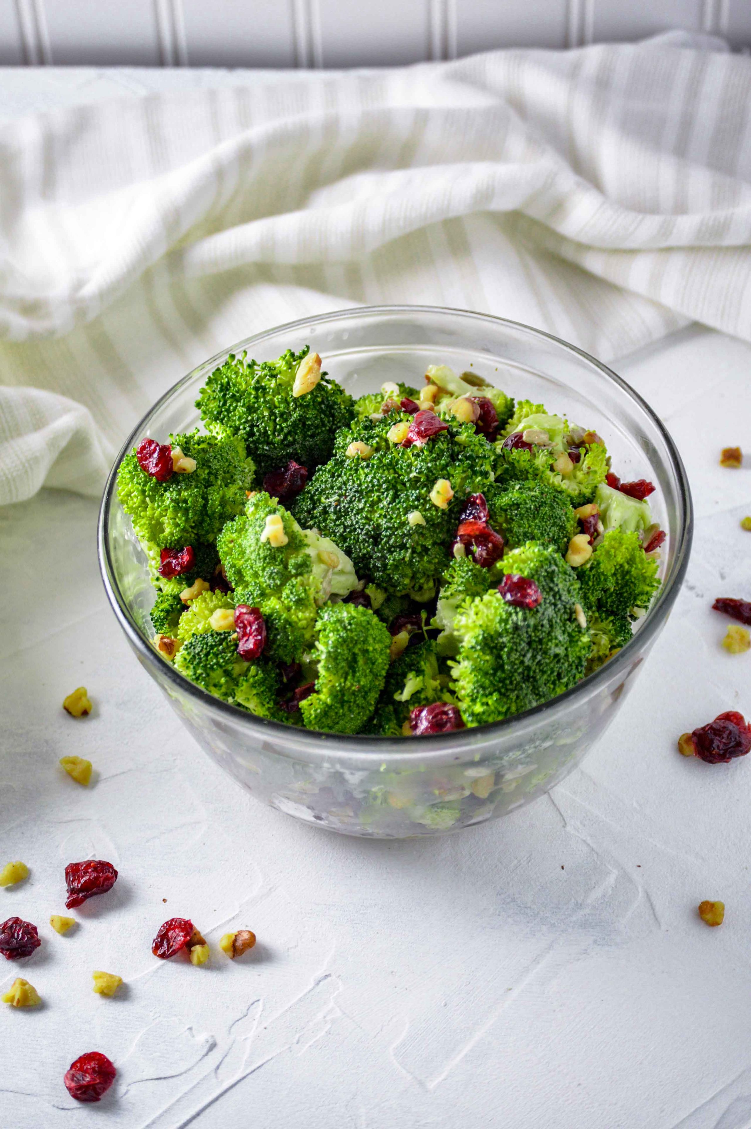 Close up top angled view of Broccoli salad in a clear bowl against a white background. there are walnuts and dried cranberries scattered around and a white towel behind the bowl of broccoli salad. https://www.atwistedplate.com/broccoli-salad/