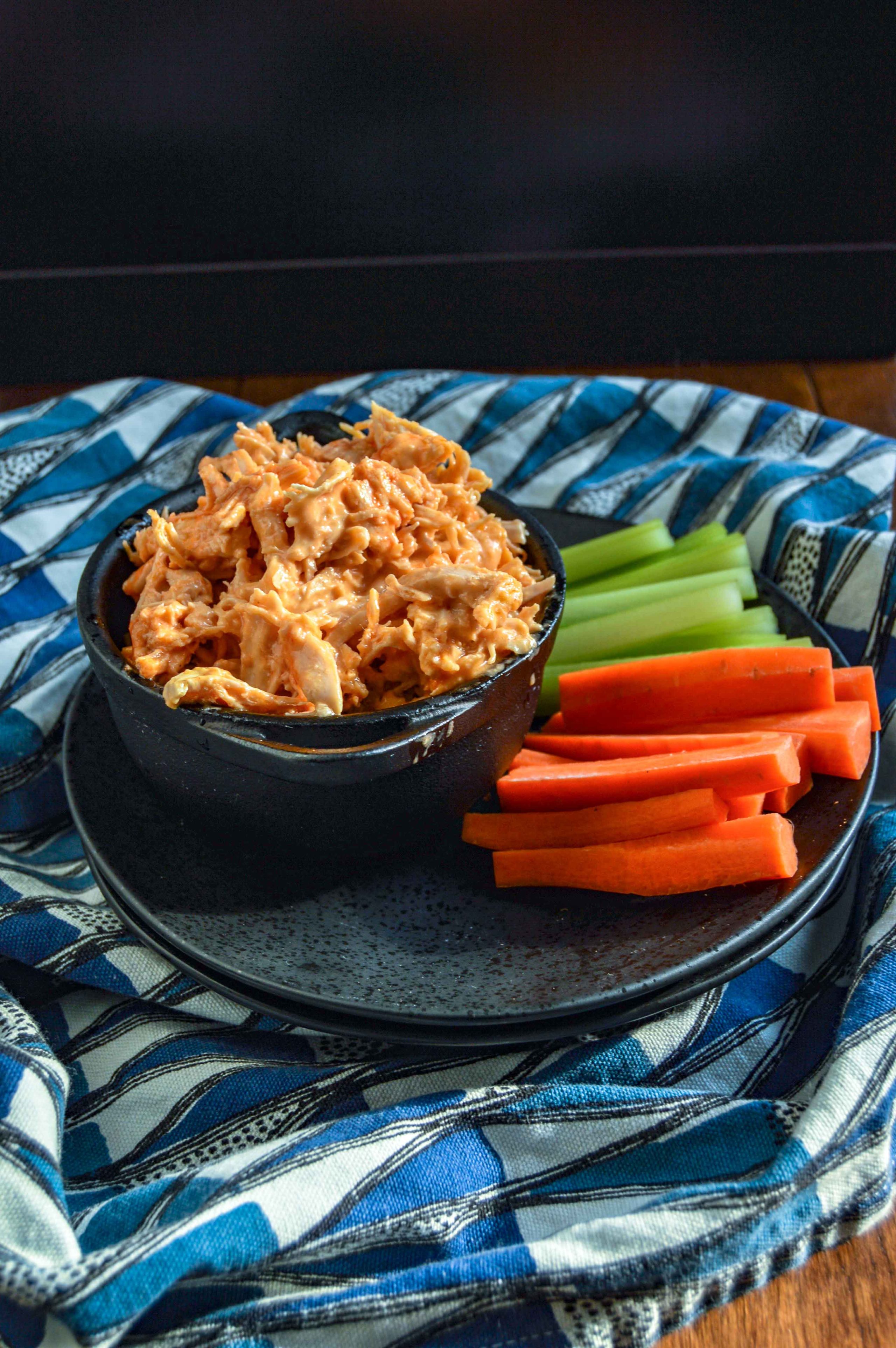 Side, slight angle view of Buffalo Chicken Dip in a black bowl on a black plate with celery and carrots. Plate is sitting on a blue and white triangle towel against a dark background. www.atwistedplate.com/buffalo-chicken-dip/