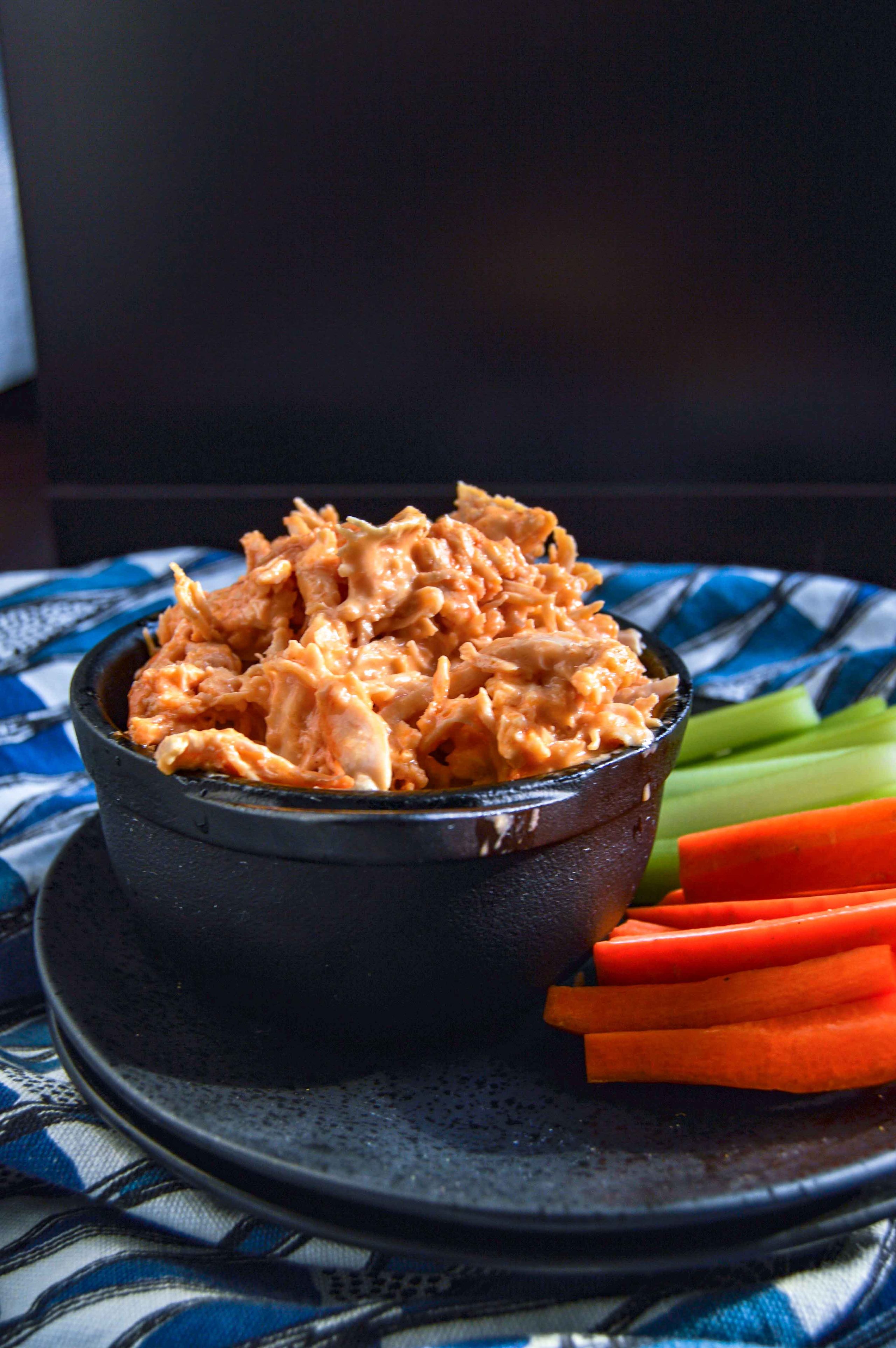 Side, slight angle view of Buffalo Chicken Dip in a black bowl on a black plate with celery and carrots. Plate is sitting on a blue and white triangle towel against a dark background. www.atwistedplate.com/buffalo-chicken-dip/