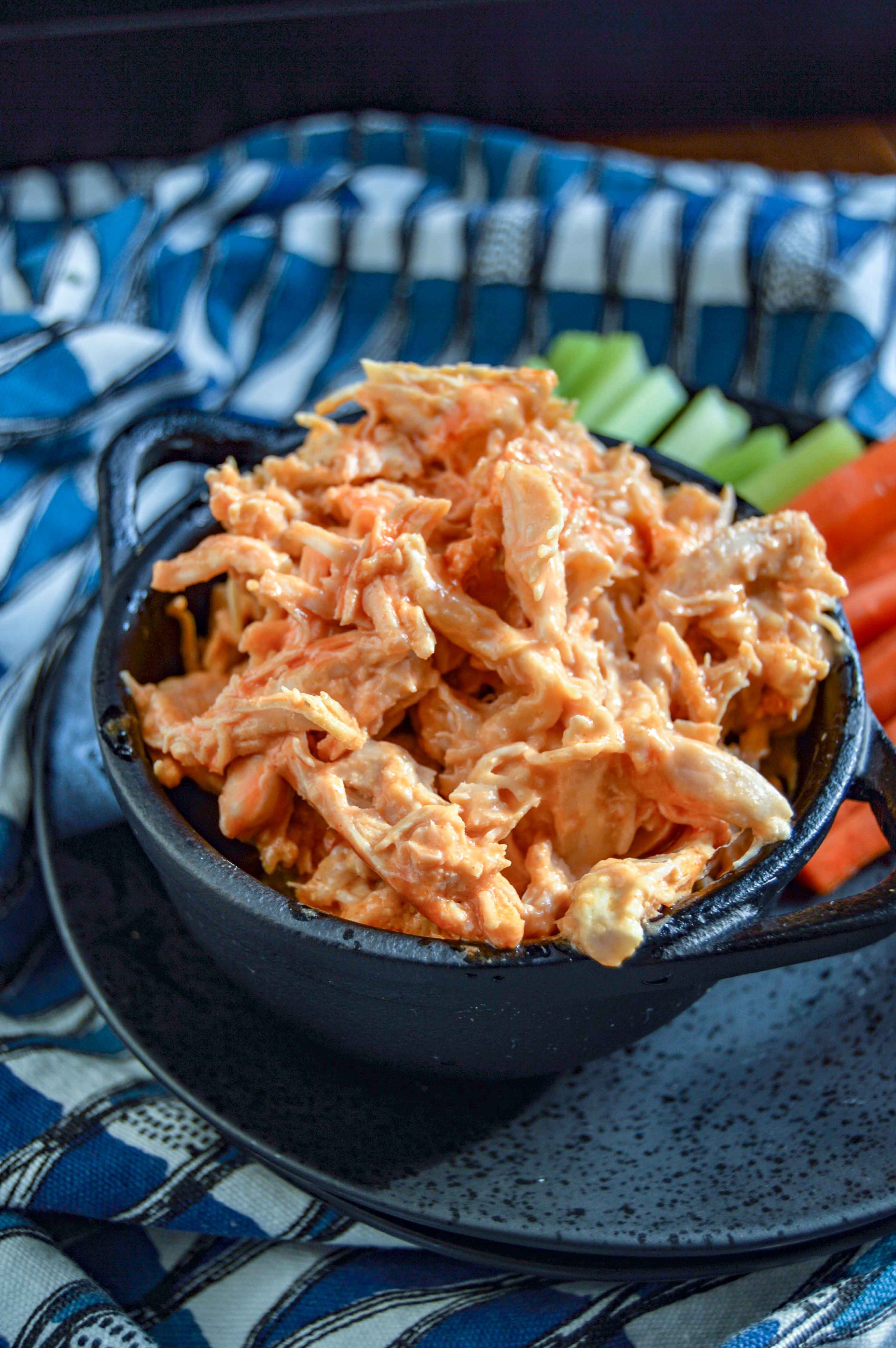 Side angle close up view of Buffalo Chicken Dip in a black bowl on a black plate with celery and carrots. Plate is sitting on a blue and white triangle towel against a dark background. www.atwistedplate.com/buffalo-chicken-dip/