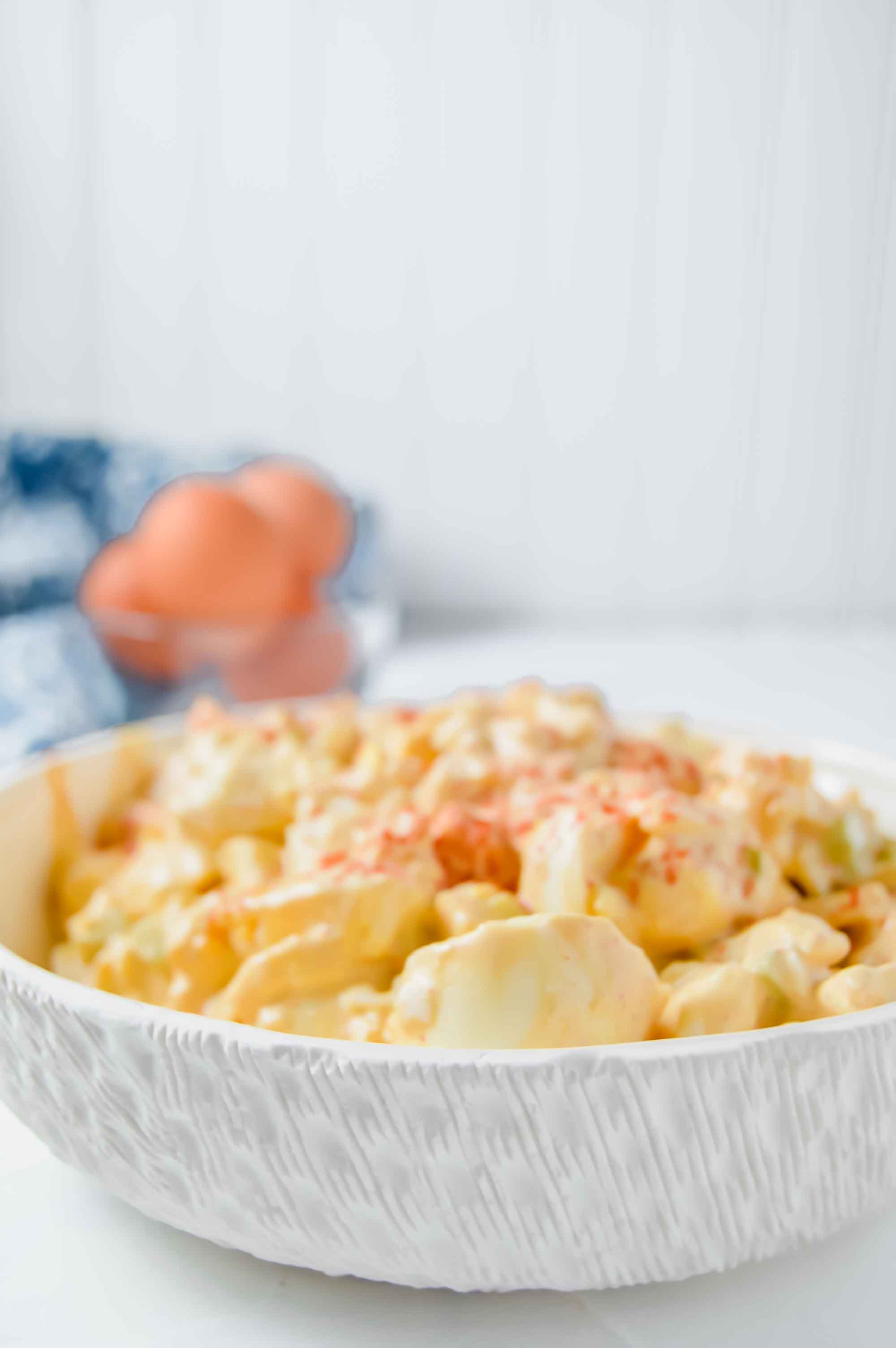 Side close up view of a white bowl with deviled egg salad. In the upper left corner there is a blurry blue and white towel with a clear bowl with 3 brown eggs that is cut off.https://www.atwistedplate.com/deviled-egg-salad/