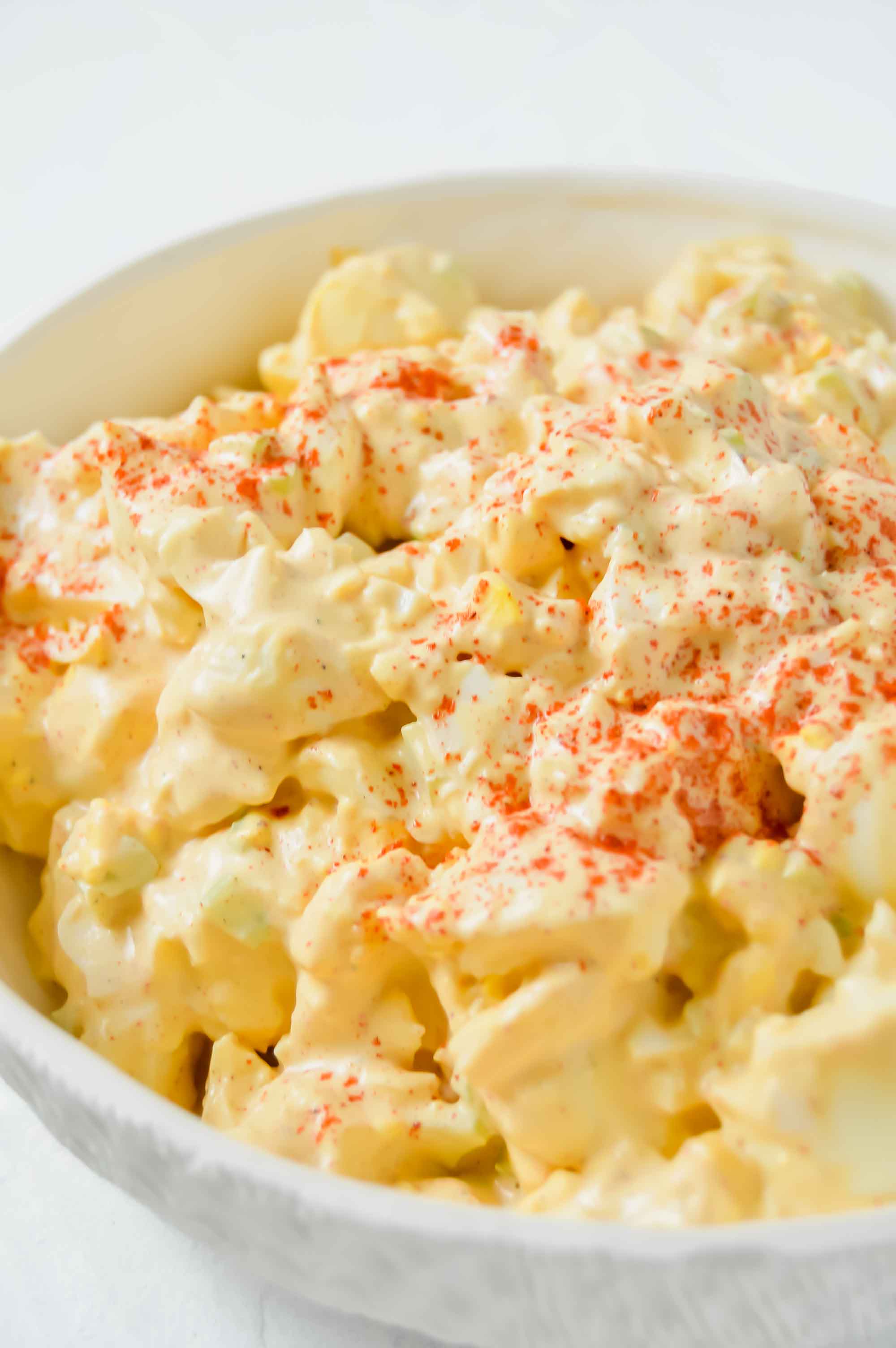 Angled close up view of a white bowl with deviled egg salad. Egg Salad has paprika sprinkled ontop. https://www.atwistedplate.com/deviled-egg-salad/