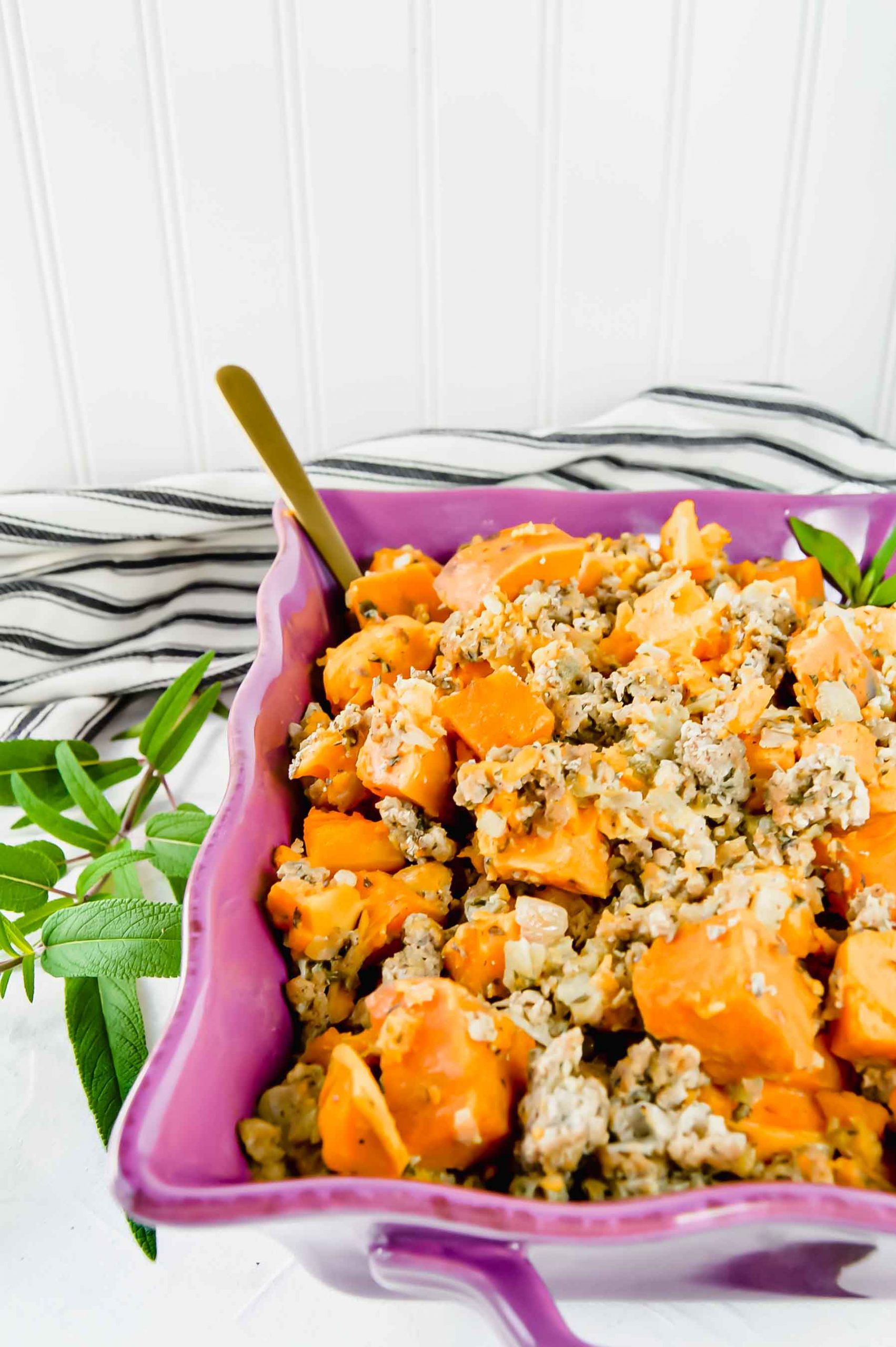 Image: angled view of Sweet Potato Stuffing in a purple dish. Around the dish are fresh sage leaves. A black and white towel is at the top of the image. https://www.atwistedplate.com/sweet-potato-stuffing/