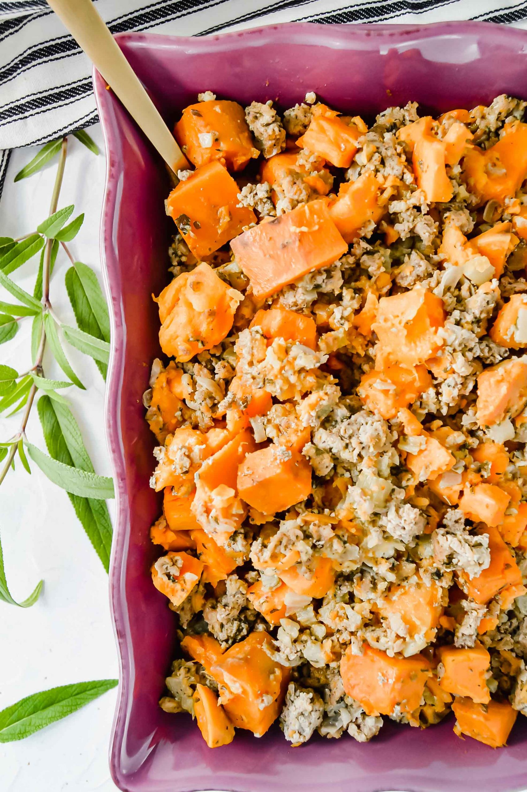 Image: Cut off top view of Sweet Potato Stuffing in a purple dish. Around the dish are fresh sage leaves. A black and white towel is at the top of the image. https://www.atwistedplate.com/sweet-potato-stuffing/