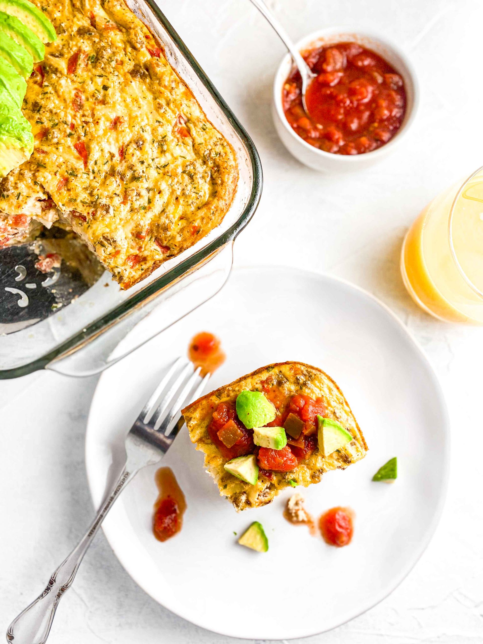 Easy Potato and Sausage Bake on a white plate topped with Avocado and Salsa with the casserole, bowl of salsa and Orange Juice against a white background. https://www.atwistedplate.com/easy-potato-and-sausage-bake/