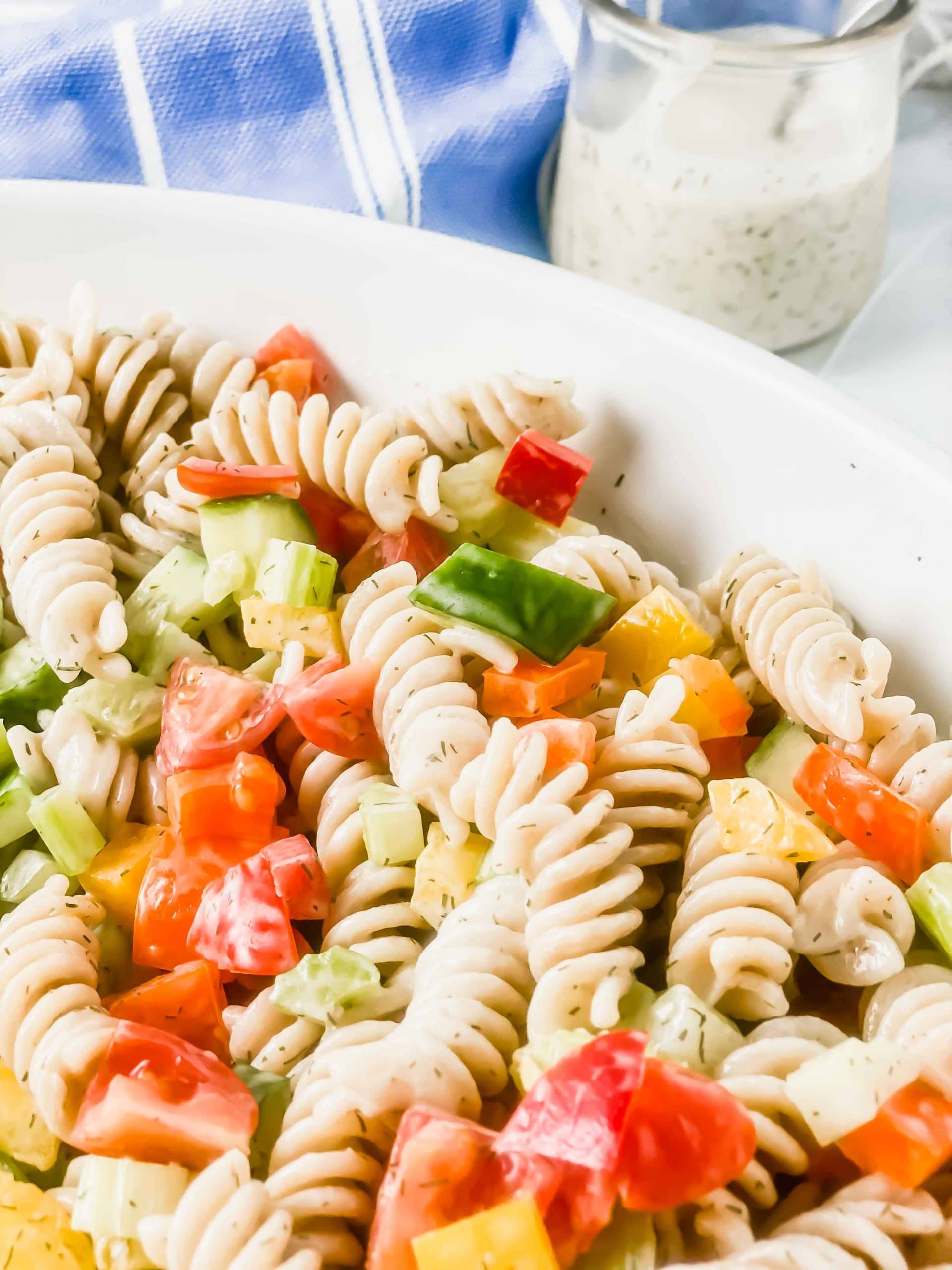 Image of Dill pasta salad in a white bowl. There is a jar of creamy dill pasta sauce and a spoon. At the top there blue bowl with a white towel. https://www.atwistedplate.com/dill-pasta-salad/