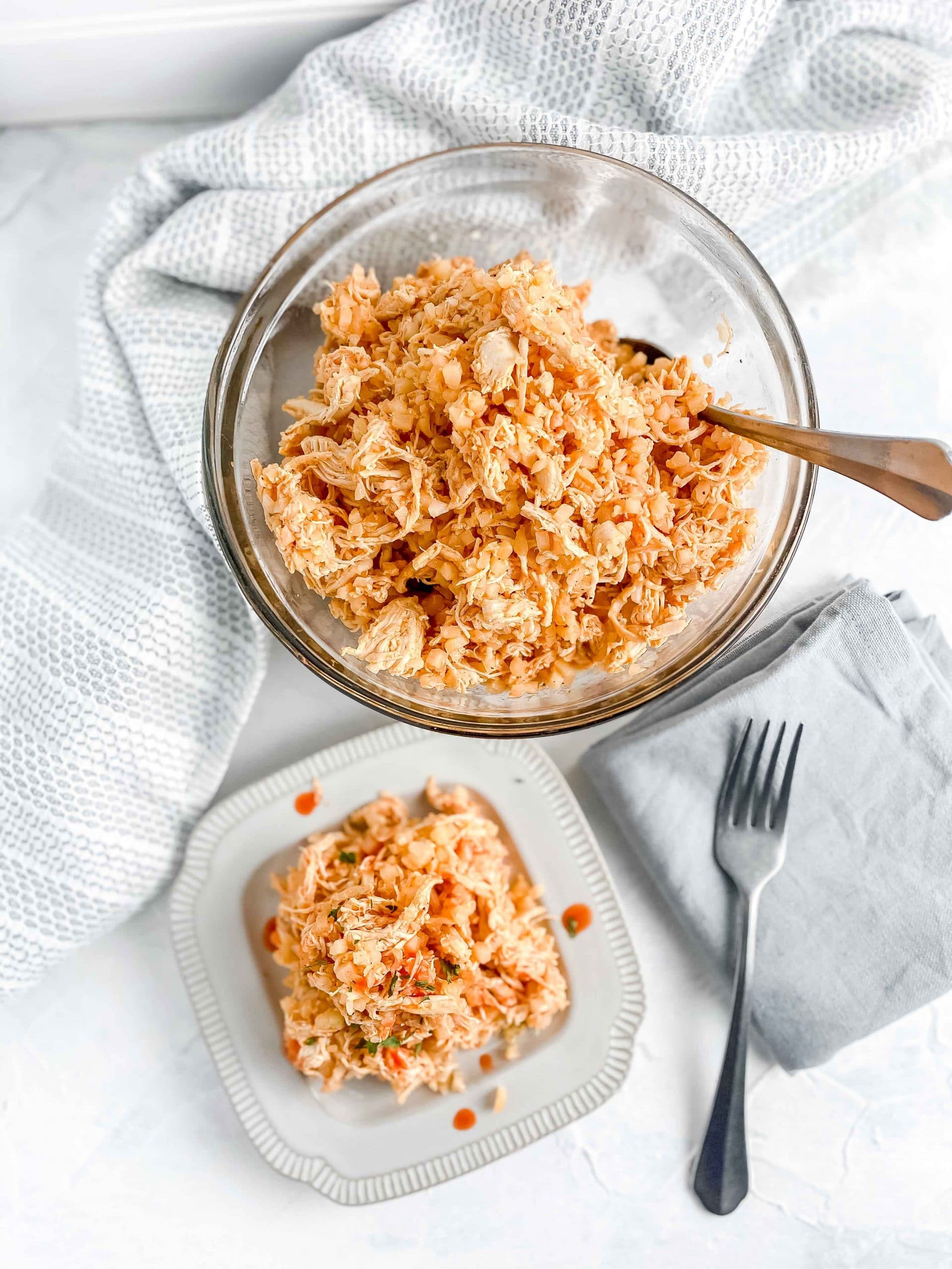 Buffalo Chicken and Cauliflower rice in a bowl with a serving on a plate topped with hot sauce. there is a fork and napkin next to the plate. https://www.atwistedplate.com/buffalo-chicken-and-cauliflower-rice/