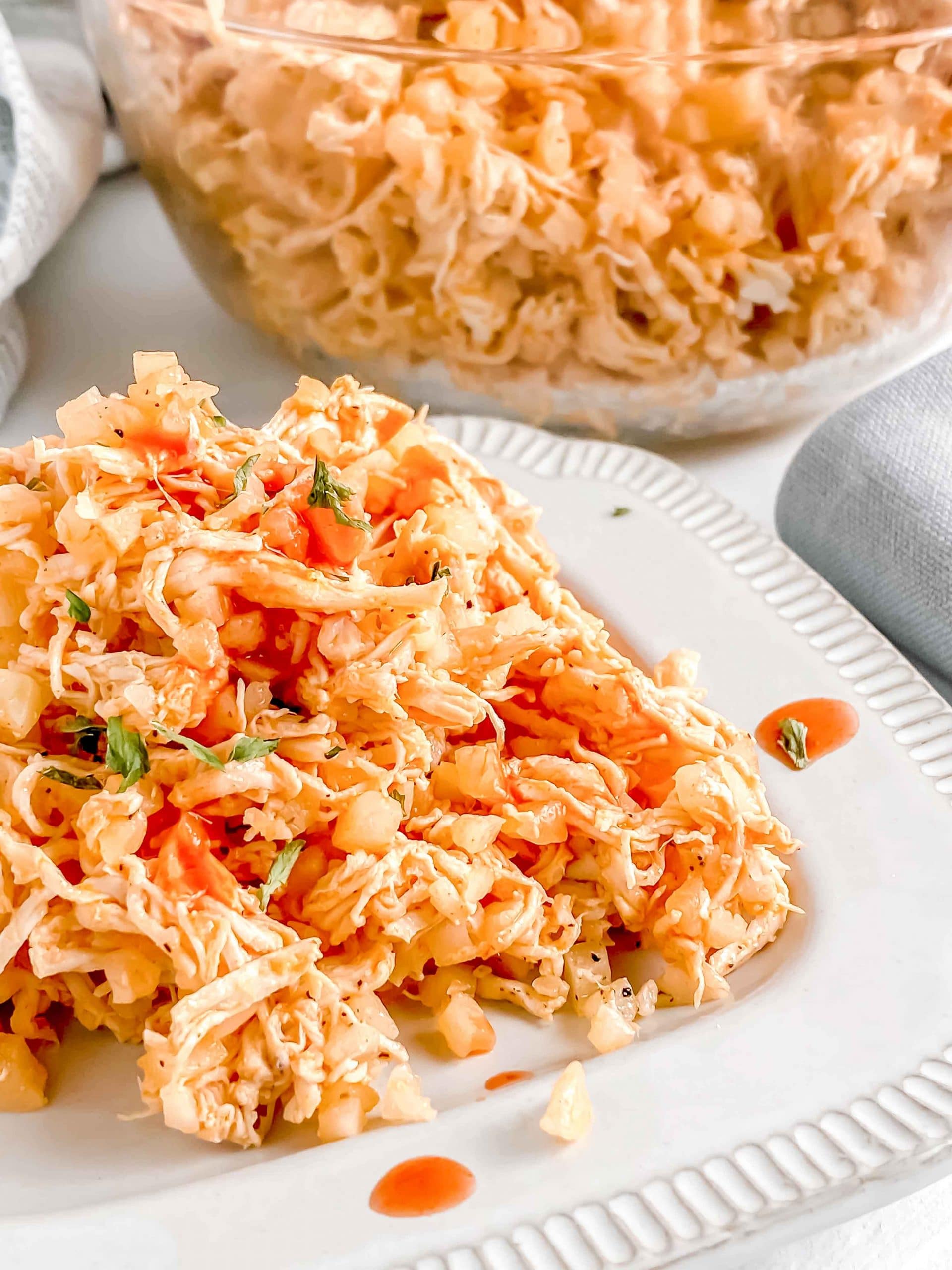Close up side view of Buffalo Chicken and Cauliflower rice in a bowl with a serving on a plate topped with hot sauce. there is a fork and napkin next to the plate. https://www.atwistedplate.com/buffalo-chicken-and-cauliflower-rice/