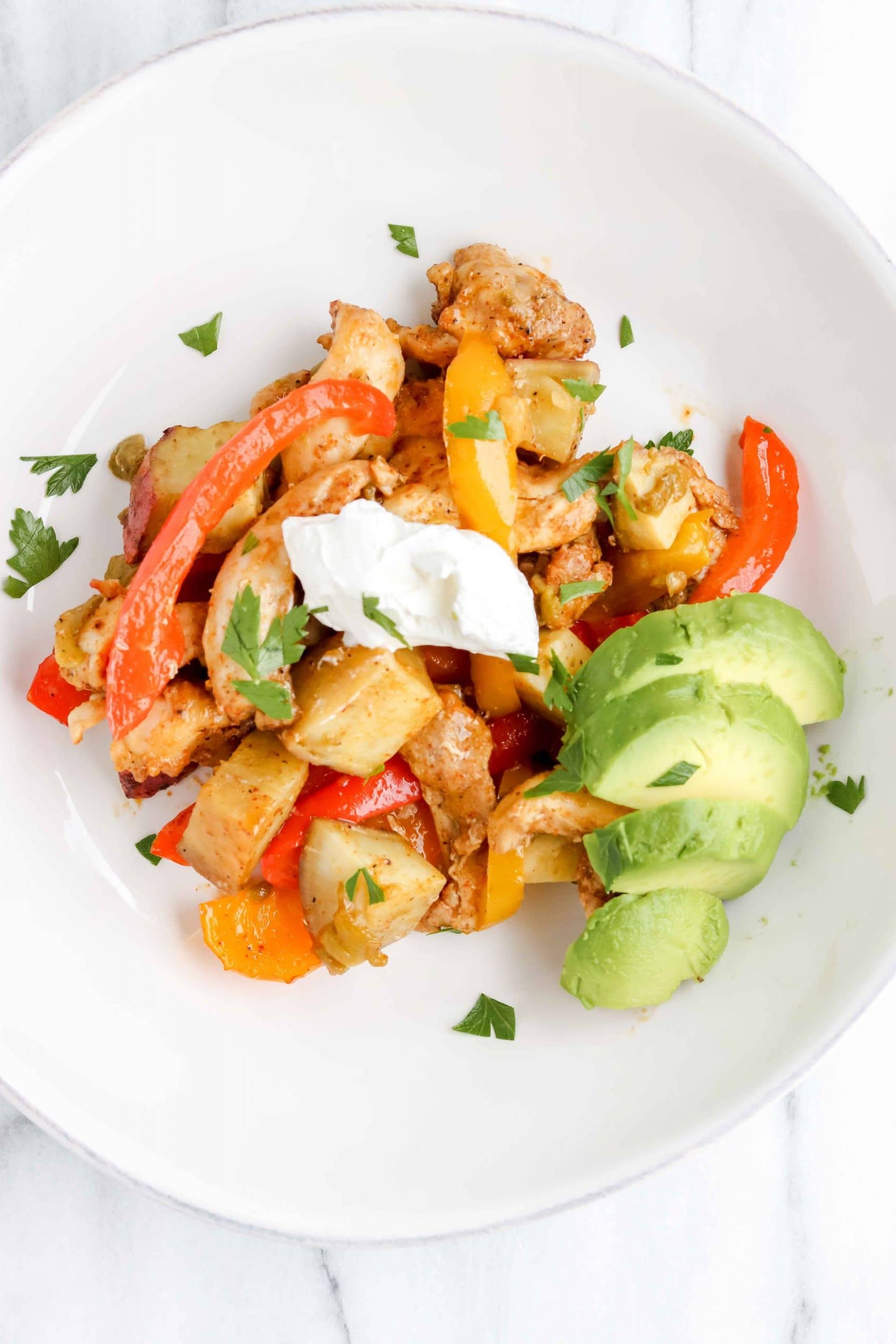 Fajita & Sweet Potato Sheet Pan Dinner in a white bowl topped with Cilantro, Sour Cream and Avocado. https://www.atwistedplate.com/fajita-sweet-potato-sheet-pan-dinner/