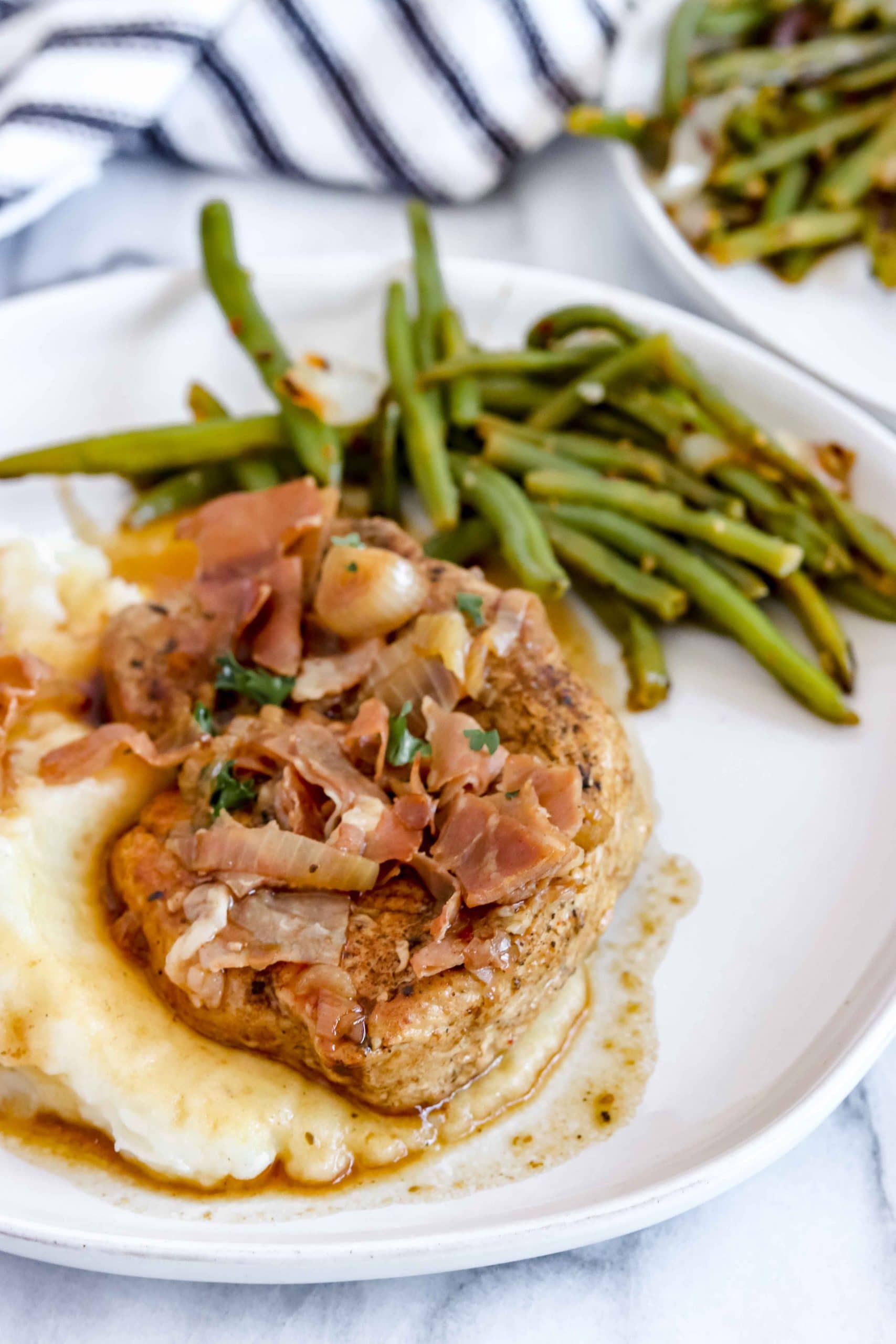 Saucy Pork Chops with Prosciutto. White round plate with Mashed Potato topped with a pork chop, prosciutto and sauce and green beans on the side of the mashed potatoes. There is a cut off plate of mashed potatoes behind it and a black and with striped towel.
www.atwistedplate/saucy-pork-chops/